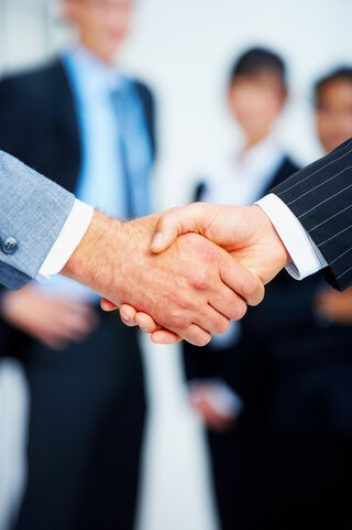 Das Bild zeigt zwei Hände, die sich in einem Handschlag treffen. Im Hintergrund sind verschwommen Menschen in Anzug und Businesskleidung zu sehen.