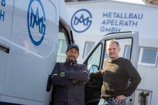 Zwei Männer stehen an einem Lieferwagen vor einem Firmengebäude mit dem Logo von Metallbau Apelrath. Sie wirken freundlich und entspannt.