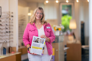 Eine lächelnde Frau in einem rosa Blazer hält eine Zeitschrift in der Hand. Im Hintergrund ist ein Geschäft mit Brillen zu sehen.