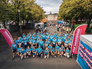 Das Bild zeigt eine große Gruppe von Läufern in blauen T-Shirts, die lächelnd posieren. Im Hintergrund sind Bäume und Veranstaltungseinrichtungen.