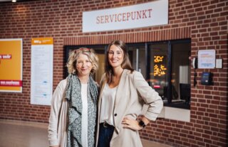 Das Bild zeigt zwei Frauen lächelnd vor einem Servicepunkt in einer modernen Einrichtung mit Backsteinwand. Sie wirken freundlich und einladend.