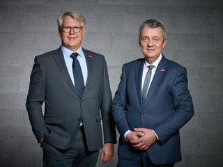 Zwei Männer in Anzügen stehen vor einer grauen Wand. Sie lächeln und wirken professionell und freundlich.