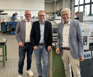 Das Bild zeigt drei Männer in einem modernen Werkstattumfeld, umgeben von Maschinen. Sie lächeln freundlich in die Kamera.