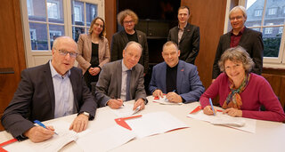Auf dem Bild sind mehrere Personen an einem Tisch versammelt, die einen Vertrag unterzeichnen. Die Atmosphäre wirkt formal und kooperativ.