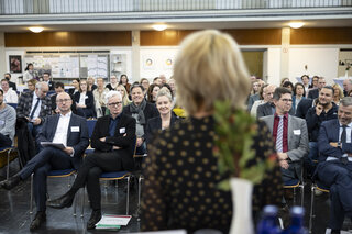Das Bild zeigt eine Konferenz mit einer Menschenmenge, die aufmerksam einer Präsentation zuschaut. Im Vordergrund ist eine Rednerin zu sehen.