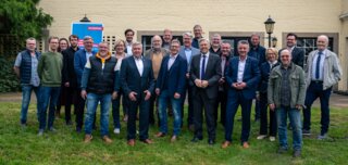 Gruppenfoto von Männern in formeller und legerer Kleidung, die vor einem Gebäude mit bunten Fenstern stehen, umgeben von Grünflächen.