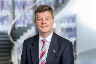 Das Bild zeigt einen Mann mit Schnurrbart in einem Anzug, der vor einer modernen Treppe posiert. Er hat einen neutralen Gesichtsausdruck.