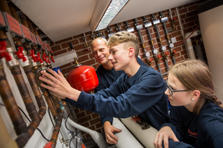 In einem Technikraum arbeiten ein Erwachsener und zwei Jugendliche gemeinsam an Rohren und Ventilen, um ein System zu warten oder zu reparieren.