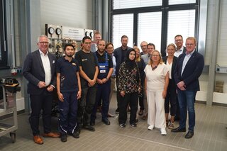 Eine Gruppe von Menschen steht in einem modernen Raum, viele lächeln. Sie scheinen an einem Projekt im technischen Bereich zu arbeiten.