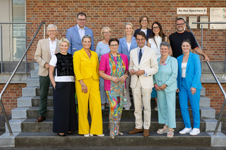 Auf der Treppe stehen 15 Personen in verschiedenen bunter Kleidung. Sie wirken freundlich und lächeln, um eine positive Atmosphäre zu erzeugen.