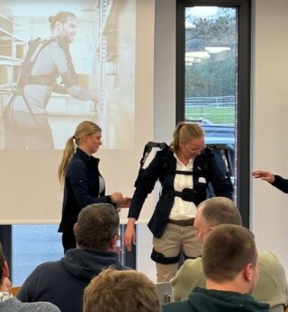 Das Bild zeigt eine Präsentation, bei der eine Person ein Exoskelett anprobiert. Im Hintergrund sieht man eine andere Person und eine Projektpräsentation.