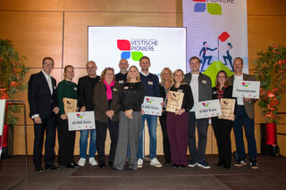 Auf dem Bild stehen mehrere Personen mit Schecks für Preisgelder. Im Hintergrund ist eine größere Anzeige mit dem Logo „Westfälische Pioniere“.