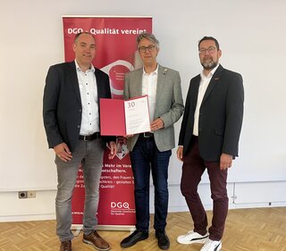 Drei Männer stehen lächelnd vor einem Banner der DGO und halten eine Urkunde. Sie scheinen eine Auszeichnung oder Anerkennung zu feiern.