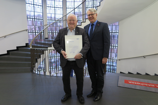 Zwei Männer stehen in einem modernen Gebäude und halten ein Zertifikat. Im Hintergrund ist eine geschwungene Treppe zu sehen.