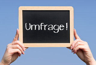 Eine Person hält eine schwarze Tafel mit der Aufschrift "Umfrage!" vor einem klaren blauen Himmel. Die Tafel hat einen Holzrahmen.