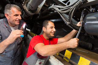 Zwei Mechaniker arbeiten unter einem Fahrzeug. Einer hält ein Werkzeug in der Hand, während der andere zuschaut und lächelt. Teamarbeit in der Werkstatt.