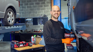Ein lächelnder Mechaniker in einer Werkstatt, der ein Werkzeug in der Hand hält. Im Hintergrund sind Autos und Werkzeuge sichtbar.
