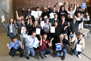 Auf dem Bild stehen viele Menschen in einem Raum, lächeln und halten Urkunden oder Dokumente in den Händen. Sie winken fröhlich.