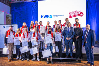 Auf dem Bild sind Absolventen mit Urkunden und roten Schals zu sehen. Sie posieren mit Vertretern der Handwerkskammer auf einer Bühne.