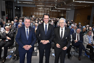 Das Bild zeigt drei Männer in Anzug, die in der Mitte stehen, umgeben von einer großen, anwesenden Menschenmenge in einem modernen Veranstaltungsraum.