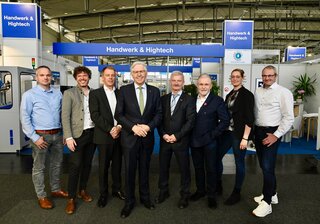 Das Bild zeigt eine Gruppe von Menschen auf einer Veranstaltung. Sie stehen zusammen vor einem Stand mit dem Thema "Handwerk & Hightech".