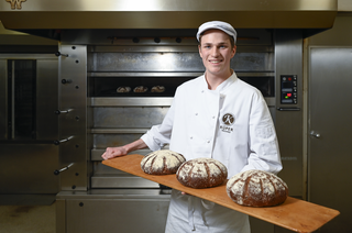 Der Bäcker in weißer Kleidung hält stolz ein Brett mit drei frischen Broten in einer Bäckerei, während im Hintergrund der Ofen sichtbar ist.