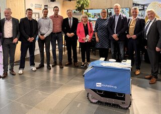 Das Bild zeigt eine Gruppe von Fachleuten und Vertretern, die gemeinsam vor einem innovativen Roboter stehen. Im Hintergrund sind Stände und Pflanzen sichtbar.