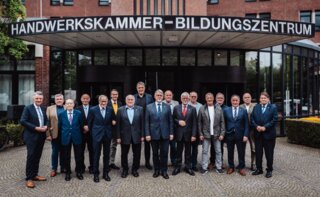 Auf dem Bild steht eine Gruppe von Männern vor einem modernen Gebäude mit dem Schild „Handwerkskammer - Bildungszentrum“. Alle lächeln freundlich.