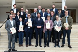 Die Gruppe steht auf einer Treppe und hält Urkunden in den Händen. Sie lächeln freundlich und sind formell gekleidet.