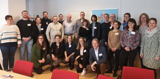 Auf dem Bild ist eine Gruppe von etwa 20 Personen in einem Raum zu sehen. Sie lächeln und stehen gemeinsam für ein Gruppenfoto.