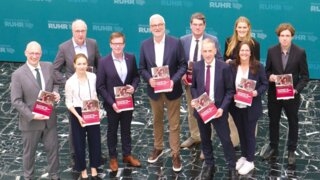 Auf dem Bild steht eine Gruppe von Personen in formeller Kleidung und hält Broschüren. Im Hintergrund sieht man ein Banner mit dem Schriftzug "Regionalverband Ruhr".