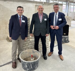 Drei Männer in Anzügen stehen in einem Bau-Workshop, umgeben von Ziegelsteinmauern und Bauwerkzeugen. Sie lächeln in die Kamera.