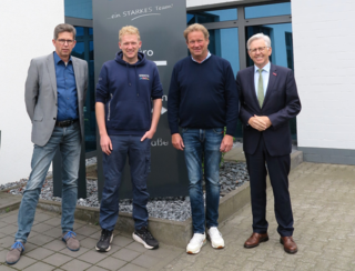 Auf dem Bild stehen vier Personen vor einem Unternehmensschild. Sie wirken freundlich und profesionell, umgeben von modernen Gebäuden.