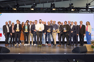 Auf der Bühne stehen zahlreiche Personen, die stolz ihre Auszeichnungen präsentieren. Eine feierliche Atmosphäre mit Dankbarkeitsbekundungen.