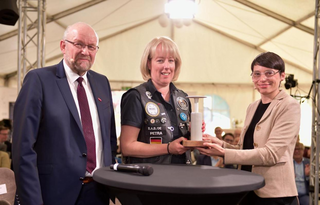 Eine Frau in einer Weste mit Abzeichen erhält einen Preis von zwei Personen. Sie stehen in einem Zelt mit Beleuchtung.