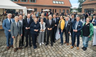 Auf dem Bild stehen mehrere Menschen in formeller Kleidung zusammen. Hintergrund: eine festliche Veranstaltung im Freien mit vielen Gästen.