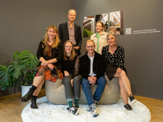 Auf dem Bild sitzen sechs Personen auf einem großen, runden Sitzmöbel. Sie lächeln und posieren vor einer grauen Wand mit zwei Bildern.