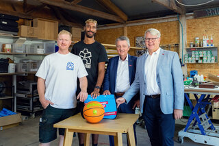 Auf dem Bild stehen vier Männer in einer Werkstatt, umgeben von Werkzeugen. Sie lächeln und halten einen Basketball sowie einen blauen Koffer.