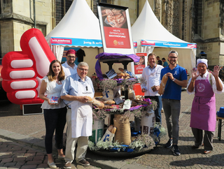 Das Bild zeigt eine Gruppe von Menschen vor einem Stand mit Brot und Backwaren. Sie lächeln, umgeben von Dekorationen und einem großen Daumen-hoch-Symbol.
