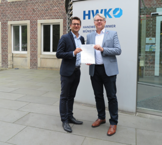 Zwei Männer stehen vor dem Gebäude der Handwerkskammer Münster, einer hält ein Dokument. Beide sind in Anzug und Hemd gekleidet.