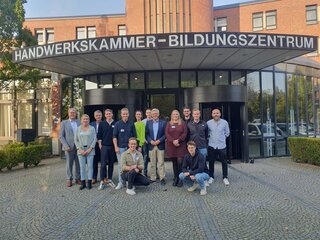 Die Gruppe steht vor dem Handwerkskammer-Bildungszentrum. Alle lächeln und wirken freundlich. Es ist ein sonniger Tag.