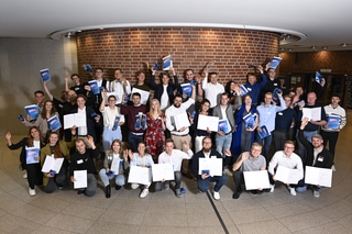 Die Gruppe von Personen steht in einem Innenraum, hält Urkunden in den Händen und lächelt. Alle sind stolz und feiern einen Erfolg.