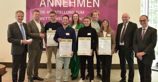 Auf dem Bild stehen mehrere Personen mit Urkunden vor einer bunten Wand. Es handelt sich um eine Preisverleihung oder Ausstellungseröffnung.